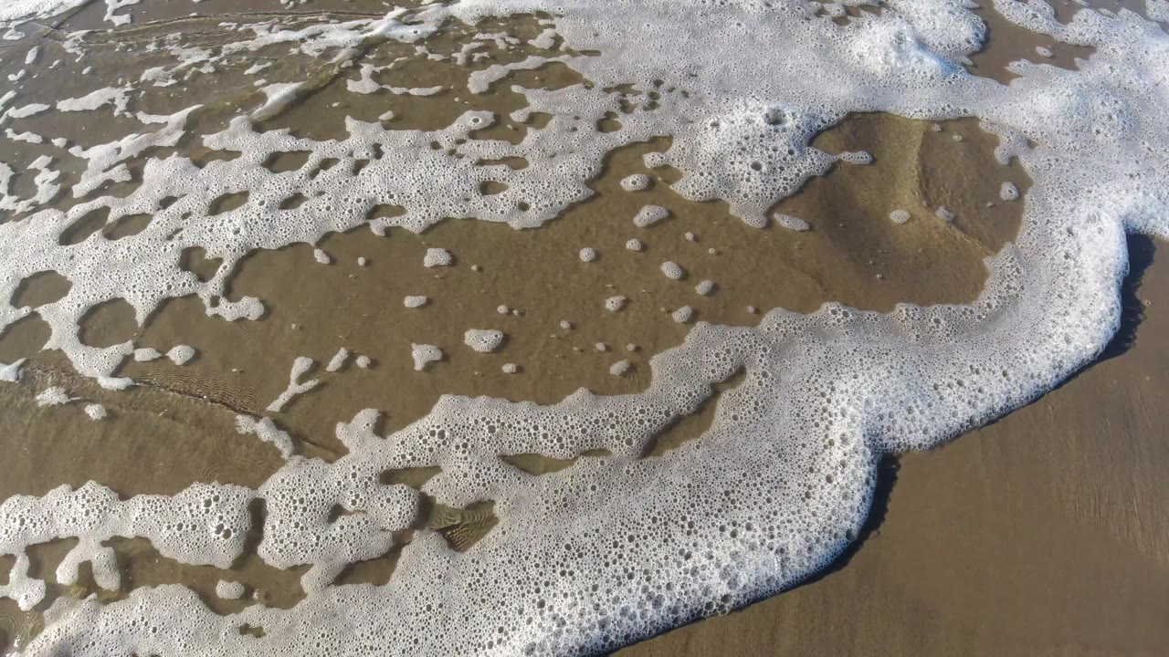 Patchy white sea foam spreads across reflective wet sand at Pismo Beach, California, with the shoreline filling the frame and no directly visible person.