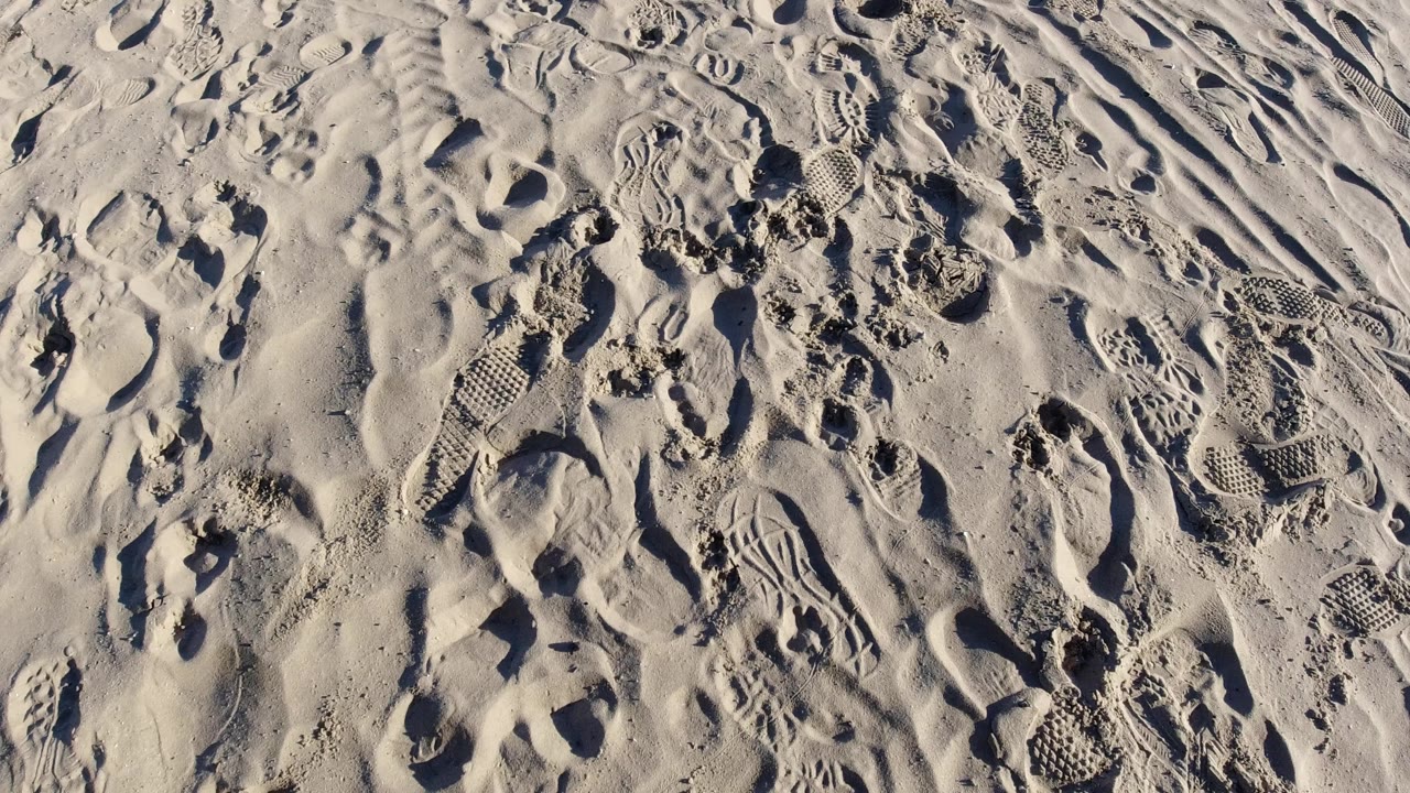 Close overhead view of Pismo Beach sand with overlapping footprints, curved shoe tracks, raised ridges, and no visible people.