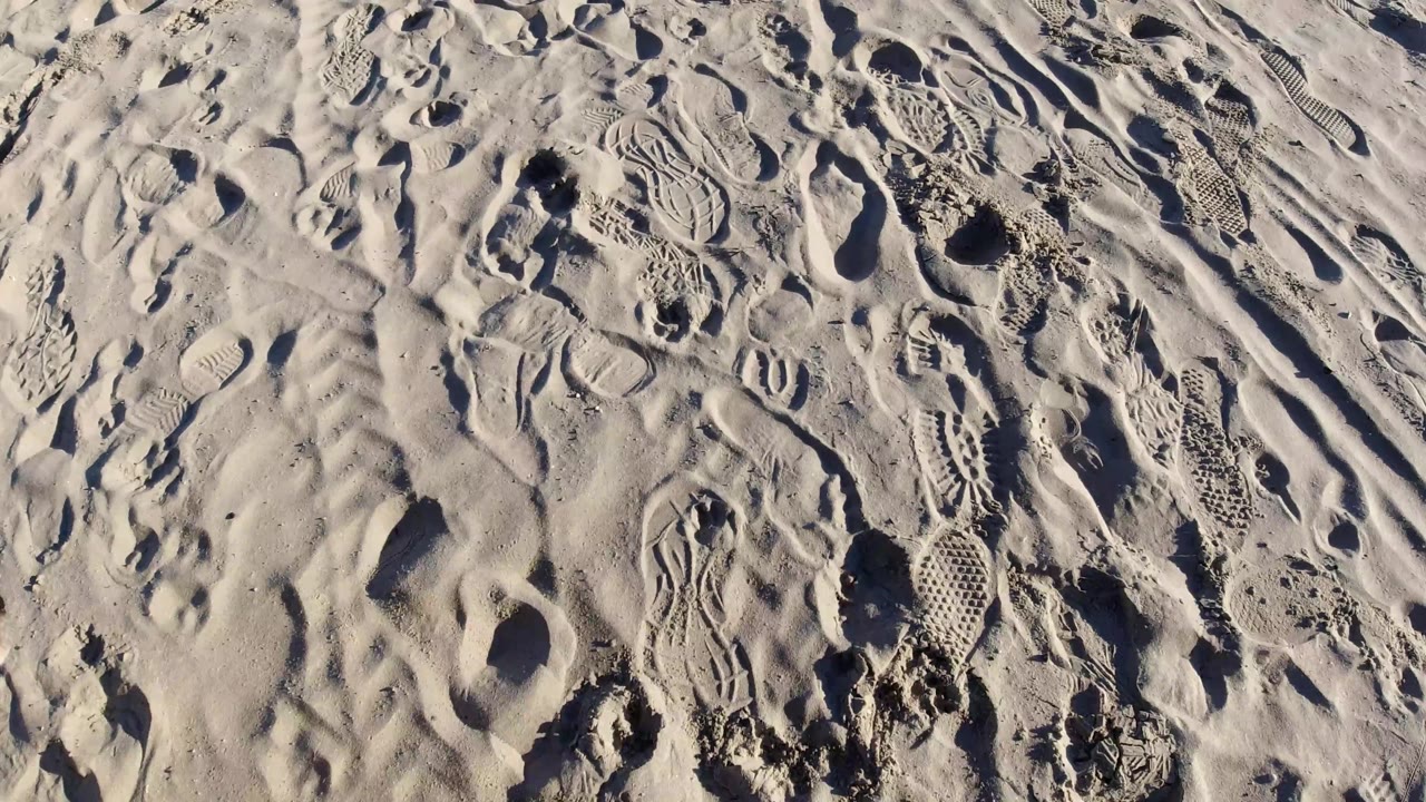 Overhead sand detail in Pismo Beach showing deeper footprint holes, diagonal tread impressions, compacted clumps, and no visible people.