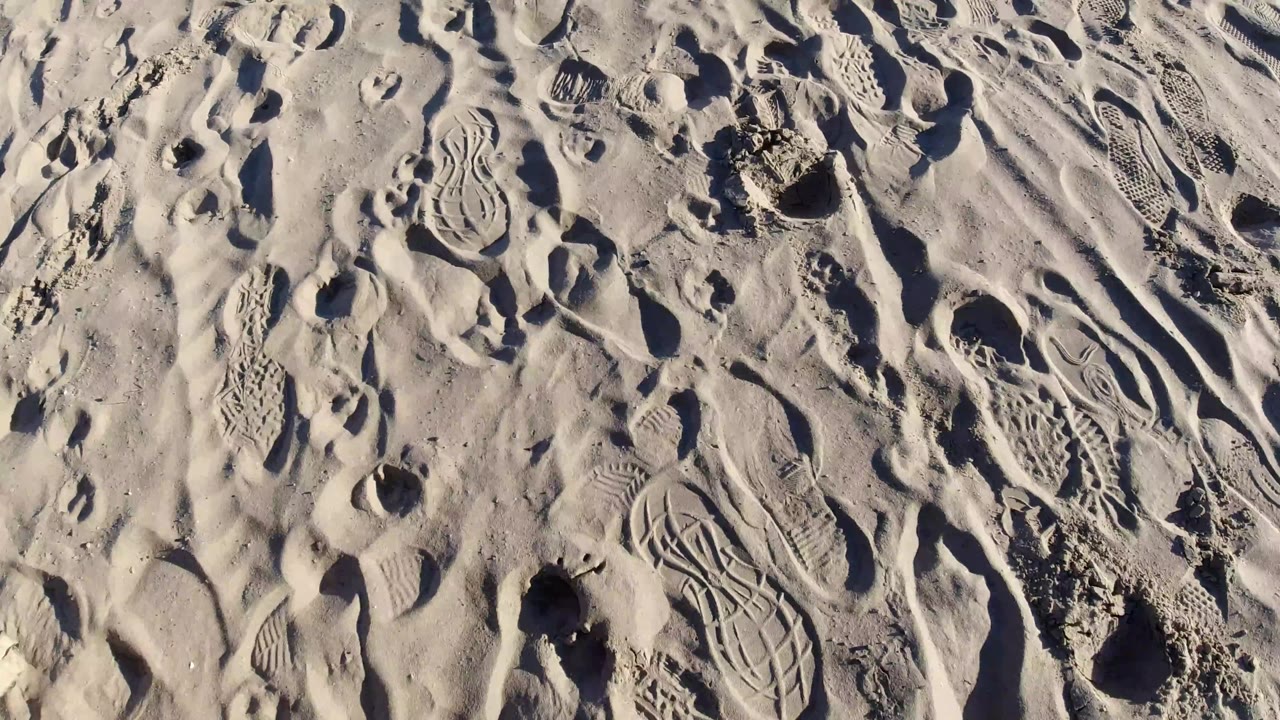 Pismo Beach sand seen from above with winding tread marks, scattered heel prints, rippled ridges, and no visible people.