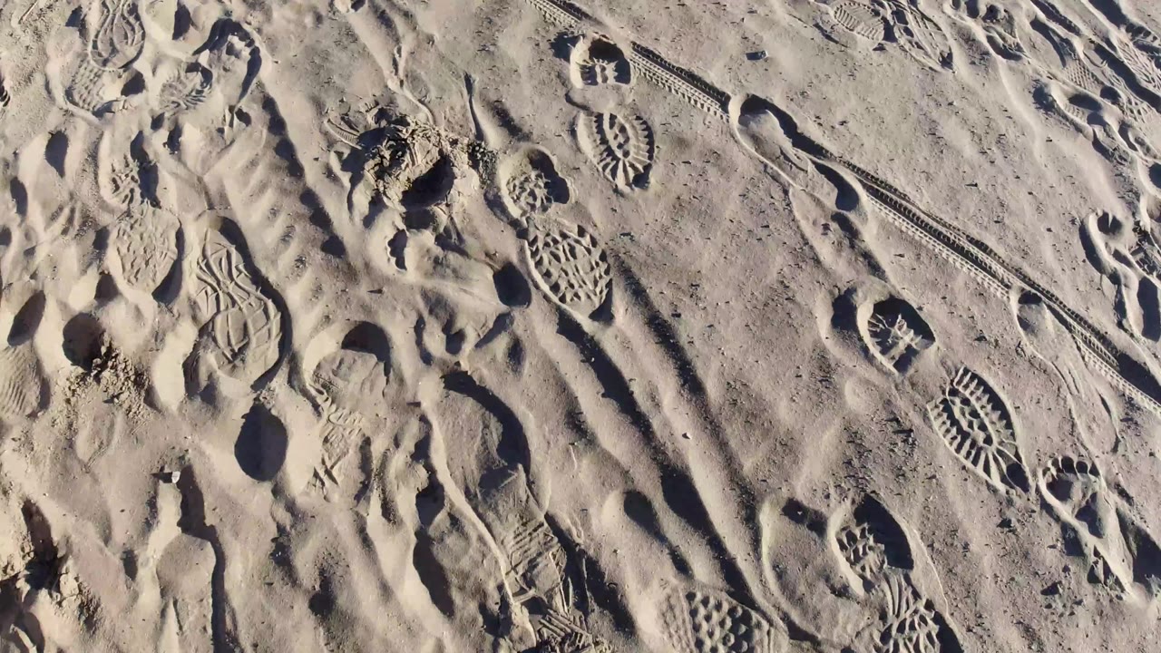 Top-down Pismo Beach sand frame with footprints, track lines, small dark clumps, and a tiny colored object near the upper left edge, with no visible people.