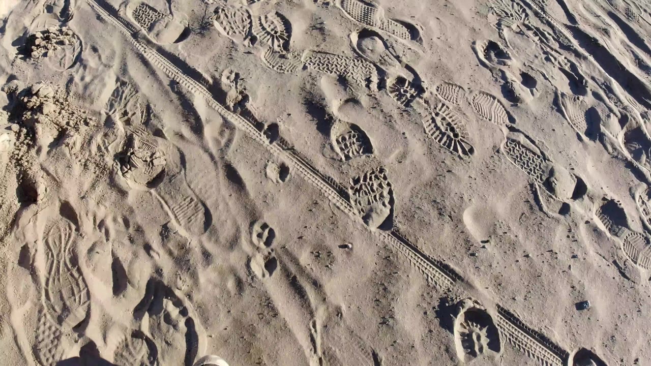 Pismo Beach sand seen from above with a diagonal track cutting across footprint patterns and raised sandy ridges, with no visible people.