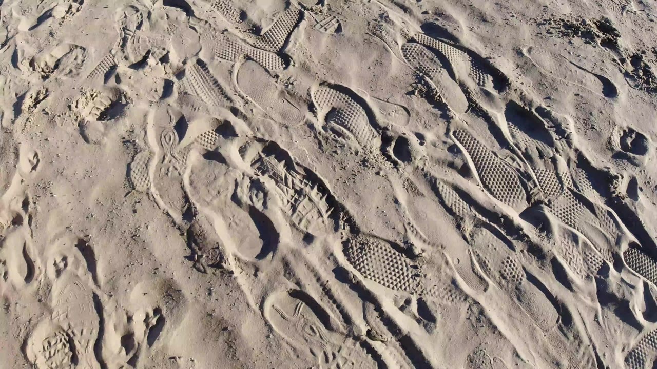 Overhead view of Pismo Beach sand covered with overlapping footprints, waffle tread impressions, ridges, and no visible people at the start of the clip.
