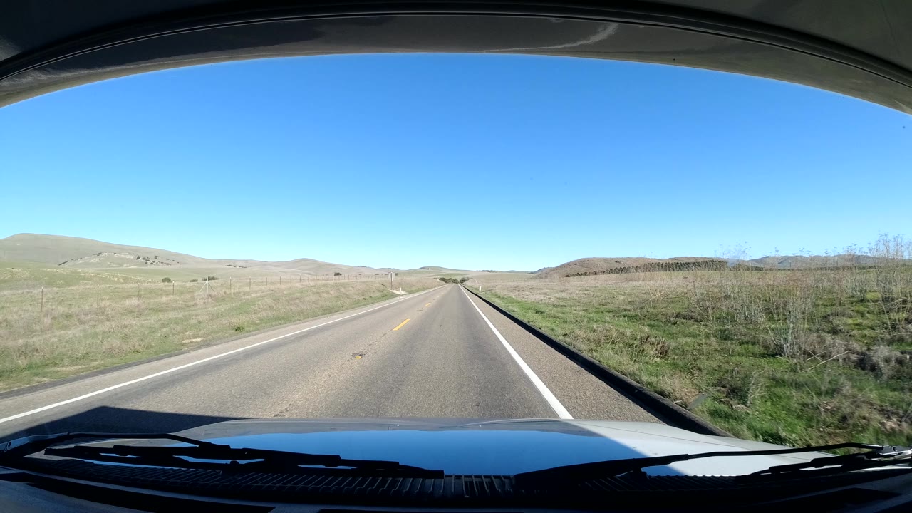 Forward windshield view on a Coast California road with rolling hills on both sides, a clear blue sky, vehicle hood and wipers at the bottom, and no visible people.