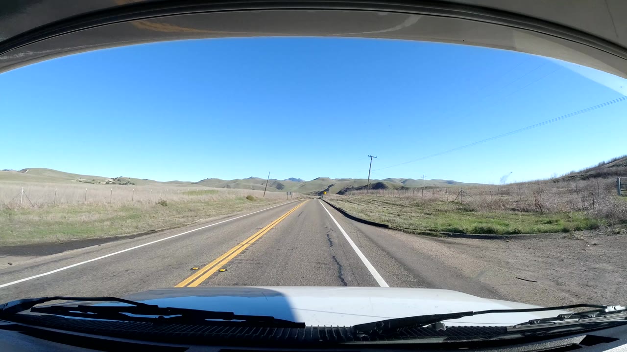 Windshield view of a Coast California two-lane road with a small roadside sign, utility poles, dry grass hills, distant ridges, and no visible people.