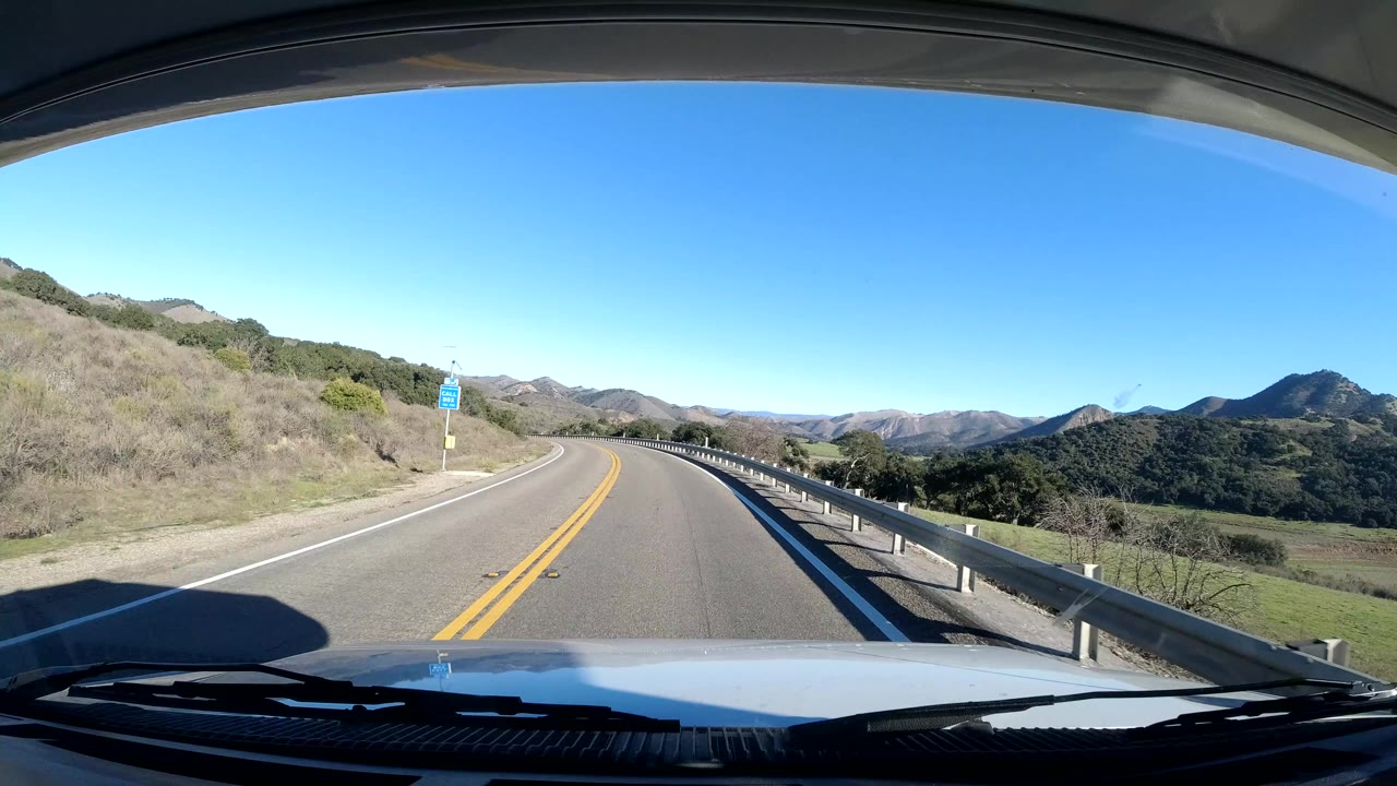 Two-lane road curving through hills with guardrail and vehicle hood visible.