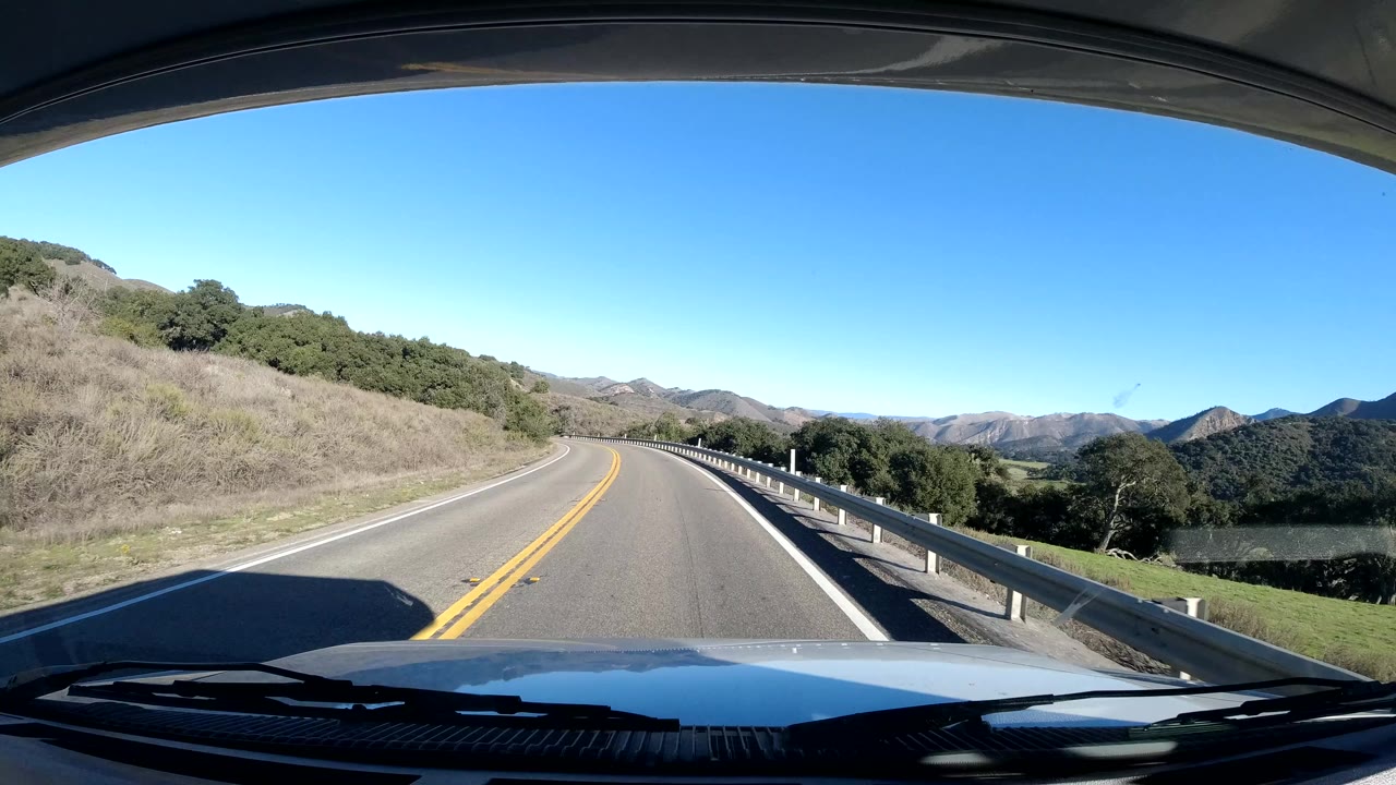 Forward vehicle view along a Coast California two-lane road, with dry brush on the left slope, guardrail on the right, mountain ridges ahead, and no visible people.