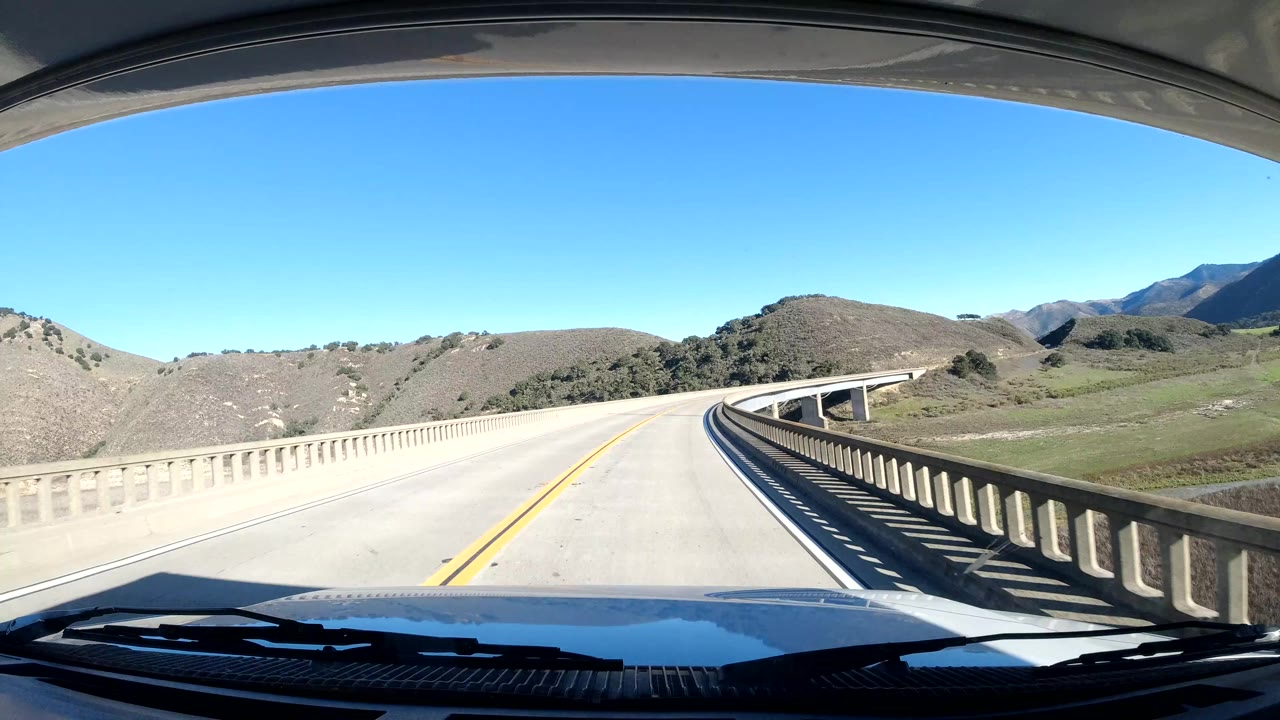 Vehicle POV crossing a concrete bridge in Coast California, with pale railings, open hills, green valley areas, distant mountains, bright sun, and no visible people.