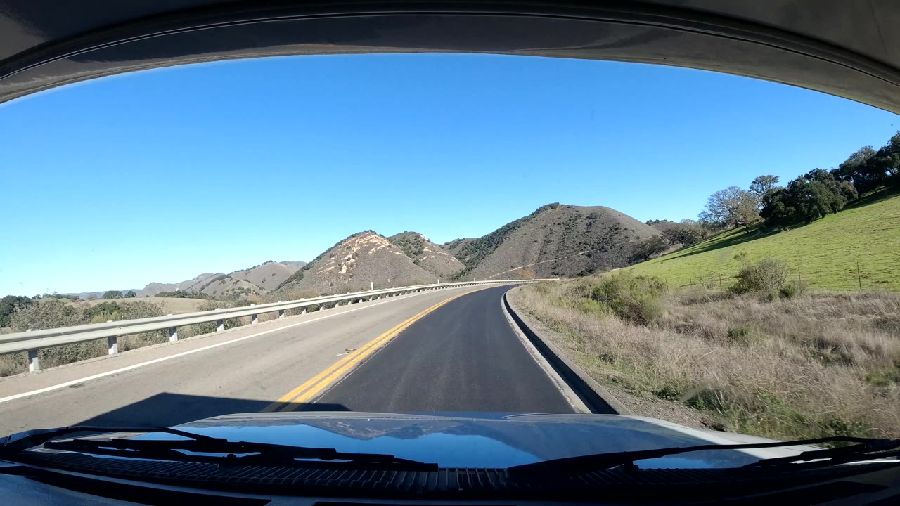 Coast California driving view on a smooth two-lane road, with bridge railings, rounded hills, a green slope to the right, clear sky, and no visible people.