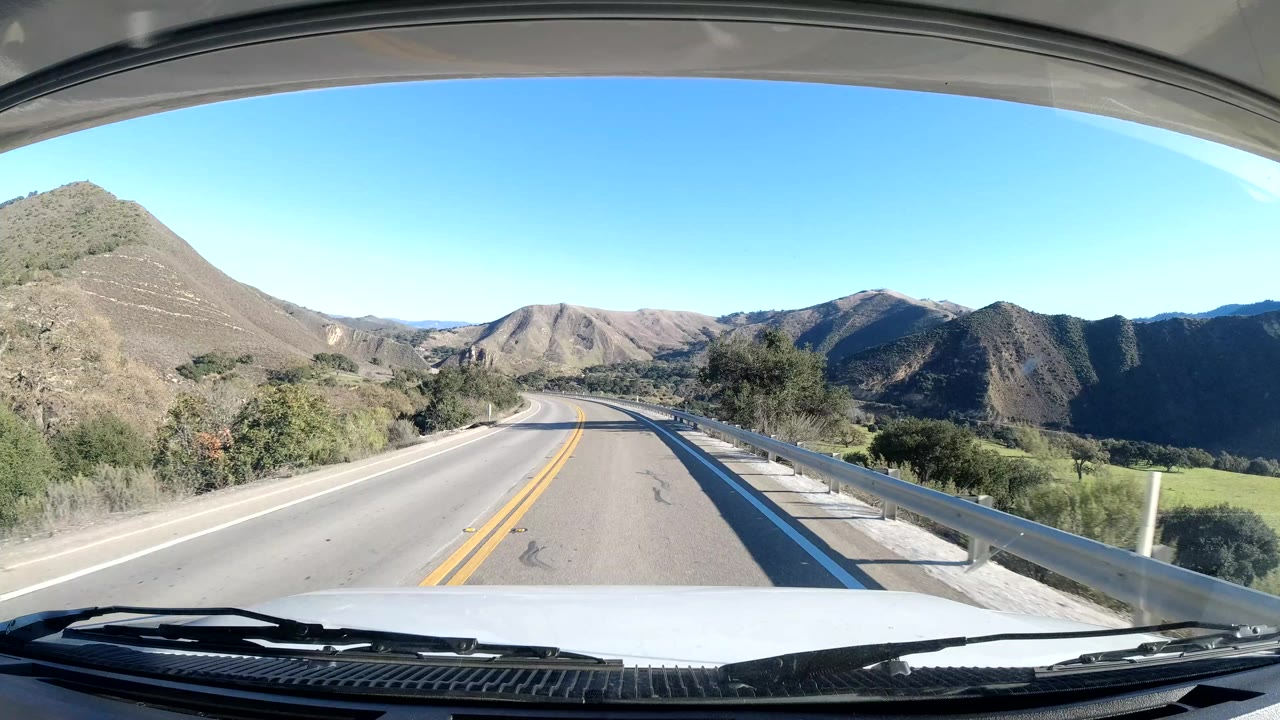 Central Coast California road view continues from the vehicle, with the lane stretching toward rounded hills, a right-side guardrail, roadside shrubs, clear blue sky, and no visible people.