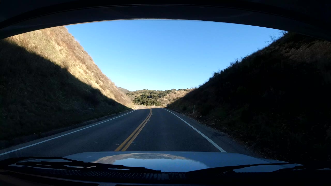 Central Coast California two-lane road passes into shadow between hillside slopes, with the vehicle hood and windshield visible, dark roadside banks, a bright sky strip, and no visible people.