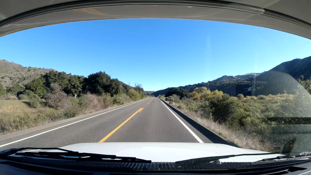 Central Coast California rural road stretches ahead from the vehicle viewpoint, with low brush along the shoulders, green hills on both sides, blue sky overhead, and no visible people.