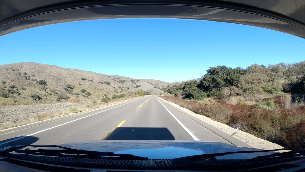 Two-lane road through dry hills seen over a vehicle hood