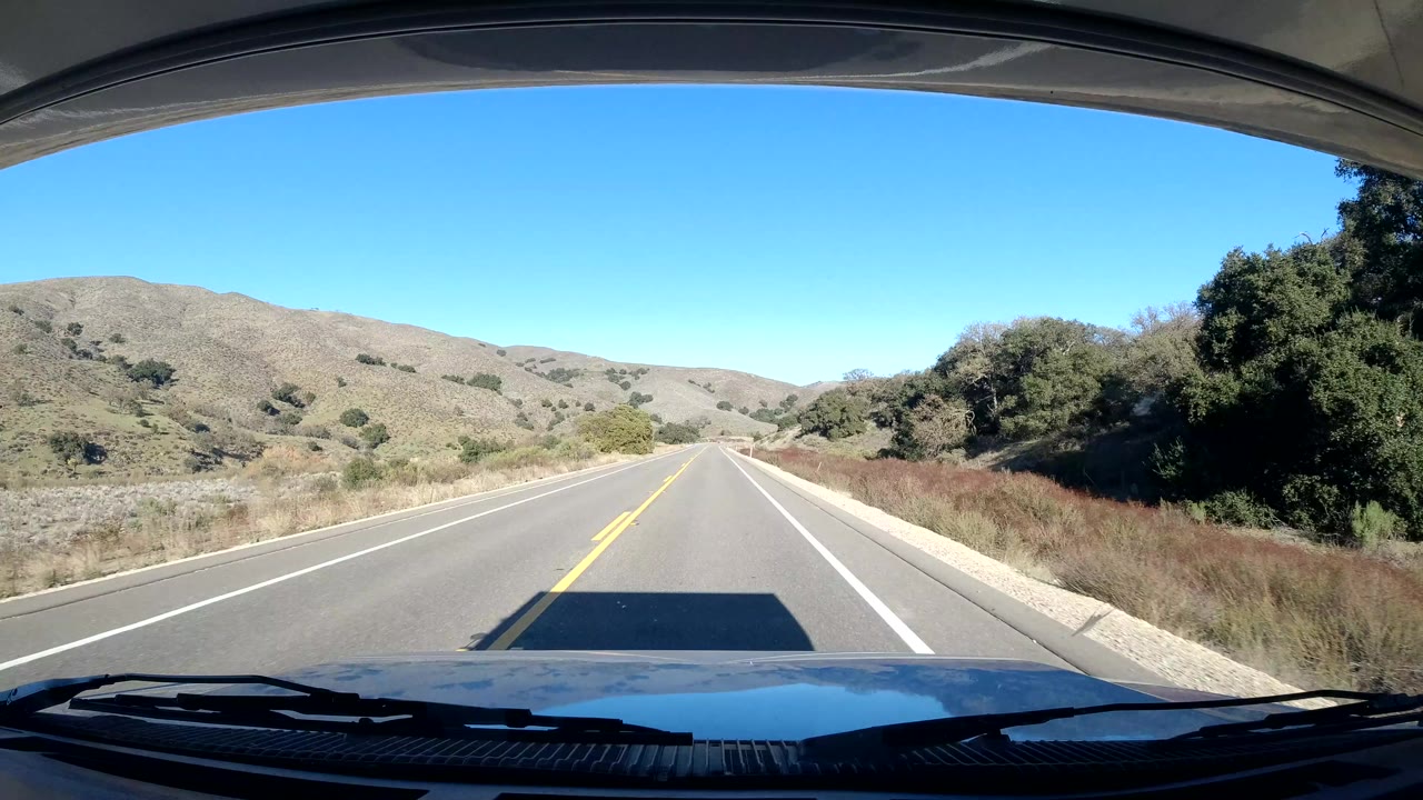 A later frame shows the road continuing straight across low Central Coast California hills, with brush along both shoulders and no visible people.