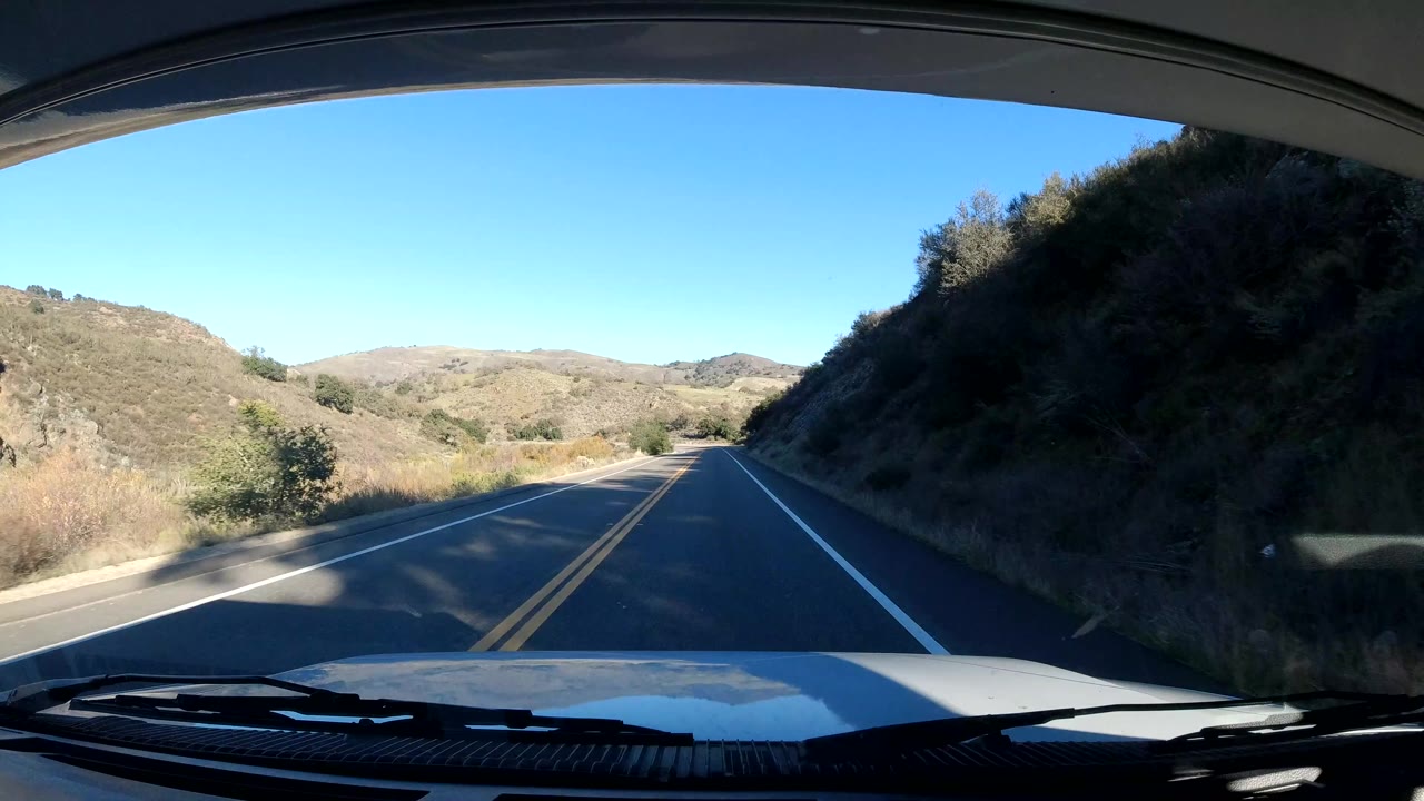 The vehicle rounds a bend on a Central Coast California road bordered by a dark hillside on the right and sunlit hills ahead, with no visible people.
