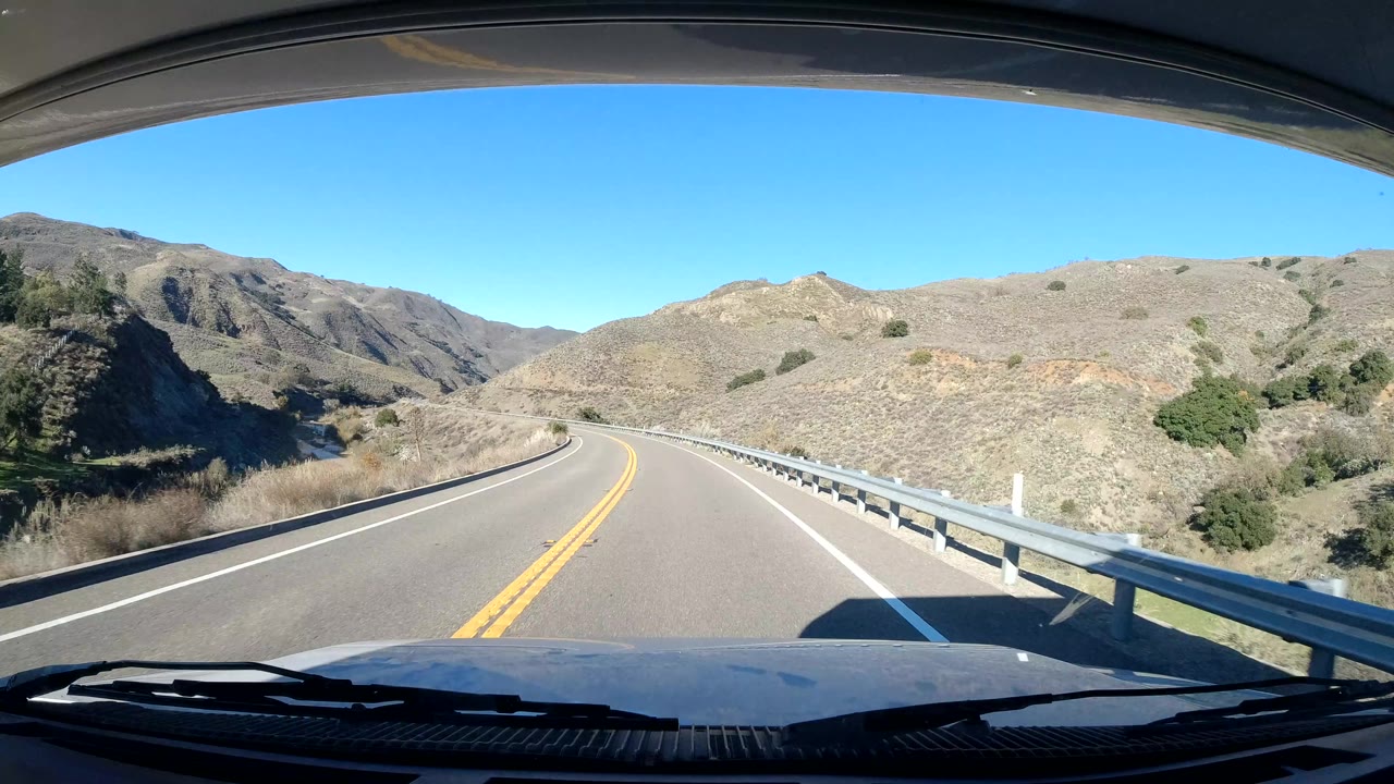 A Central Coast California road curves left past a metal guardrail, dry slopes, and rocky hills, with the vehicle hood at the bottom and no visible people.