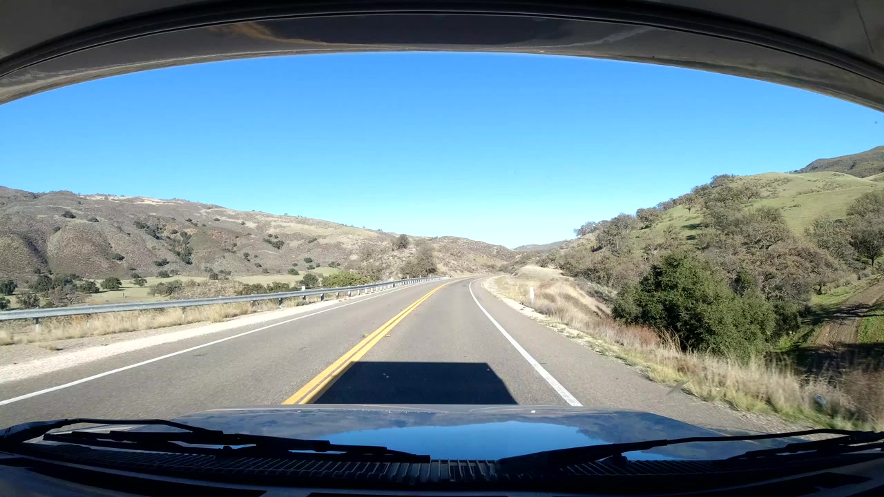 Forward view over a vehicle hood on a Coast California road, with yellow center lines, guardrail, dry hills, blue sky, and no visible people.