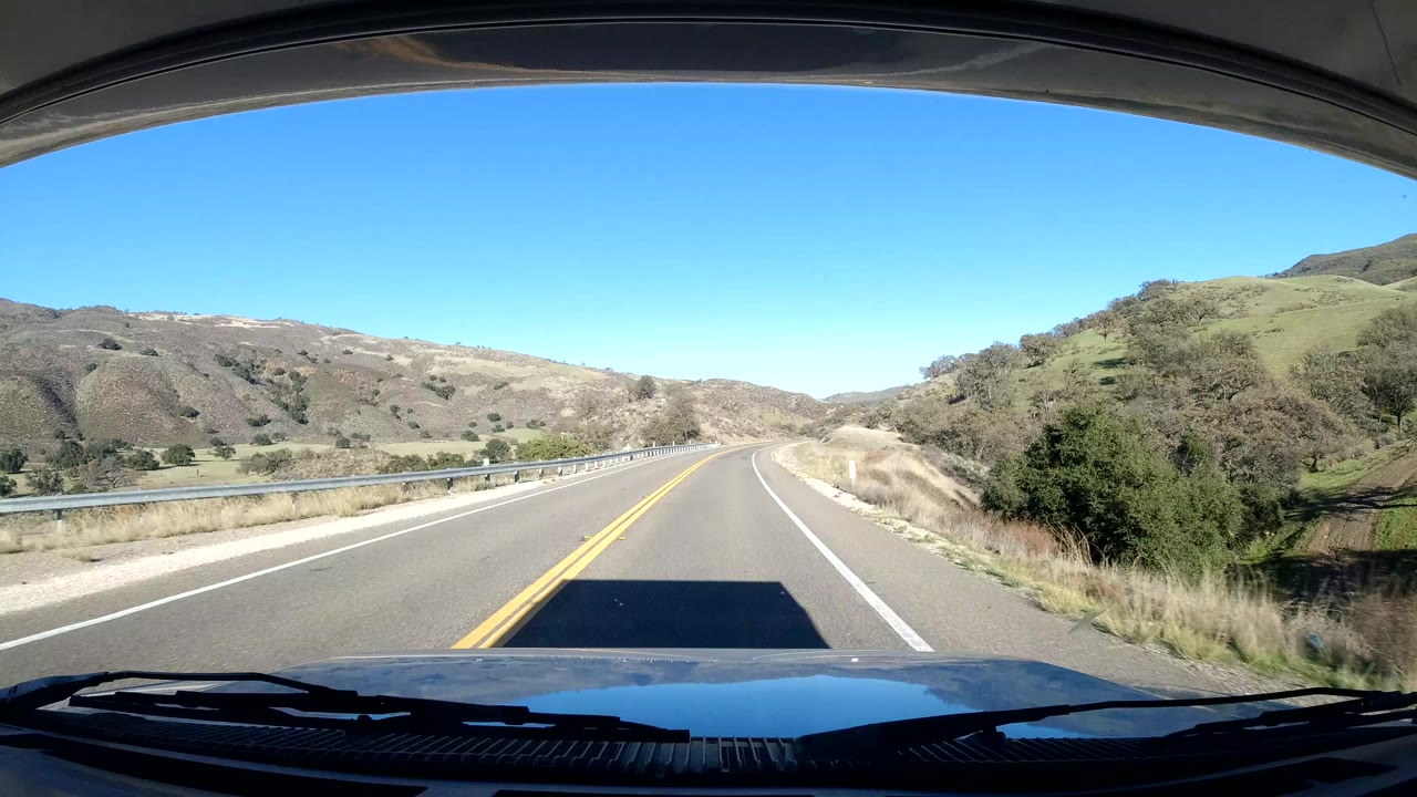 Mounted vehicle view on a Coast California road, with the lane curving slightly through dry hills, roadside guardrail, open sky, and no visible people.