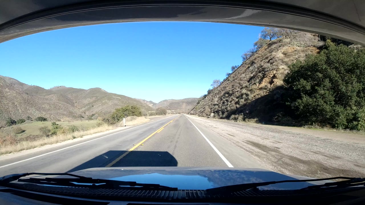 Forward road view in Coast California, with a rocky hillside close to the right shoulder, open hills ahead, vehicle hood at the bottom, and no visible people.