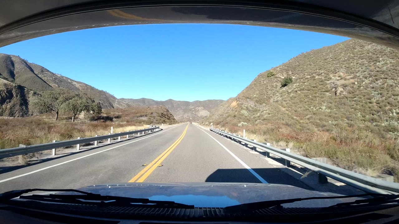 Two-lane road bordered by guardrails and low hills.