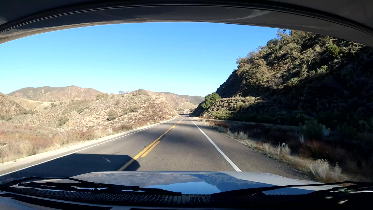 Central Coast California dash view at the start of the clip, showing a two-lane road, dry hills, blue sky, windshield edge, vehicle hood, and no visible people.