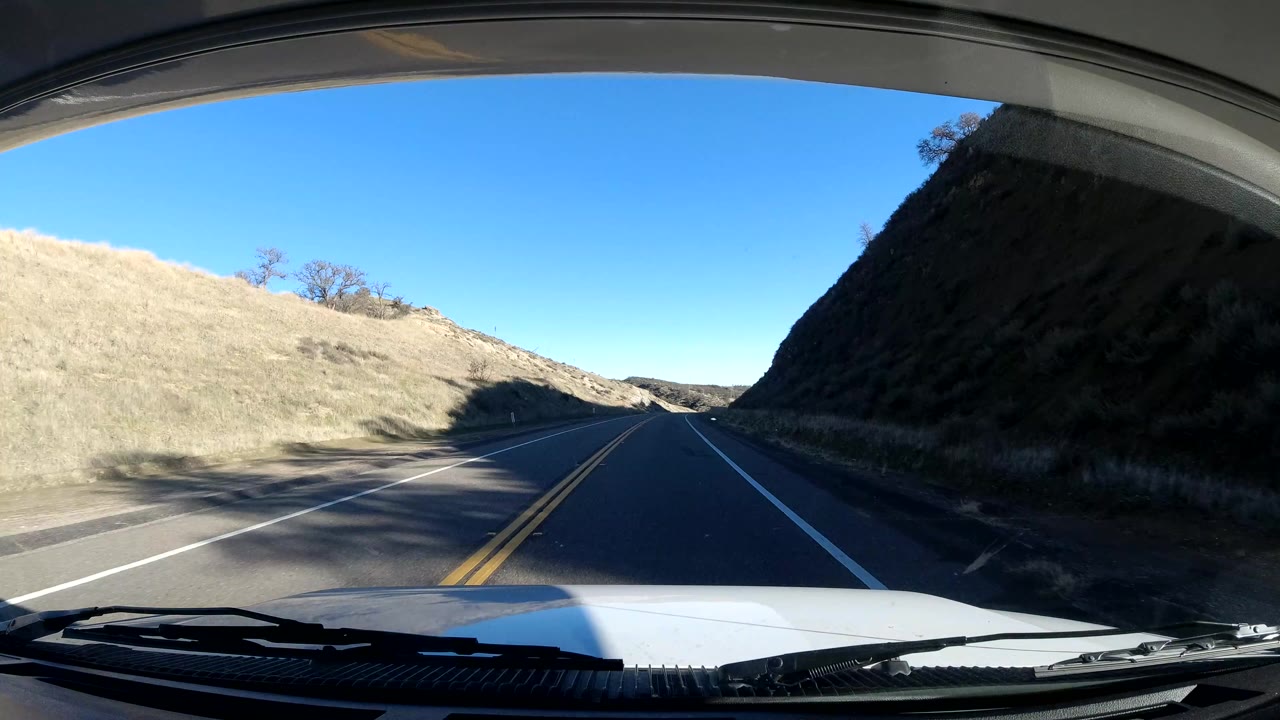 Central Coast California windshield view with the road entering a narrow stretch between a sunlit dry slope and a shadowed hill, with no visible people.