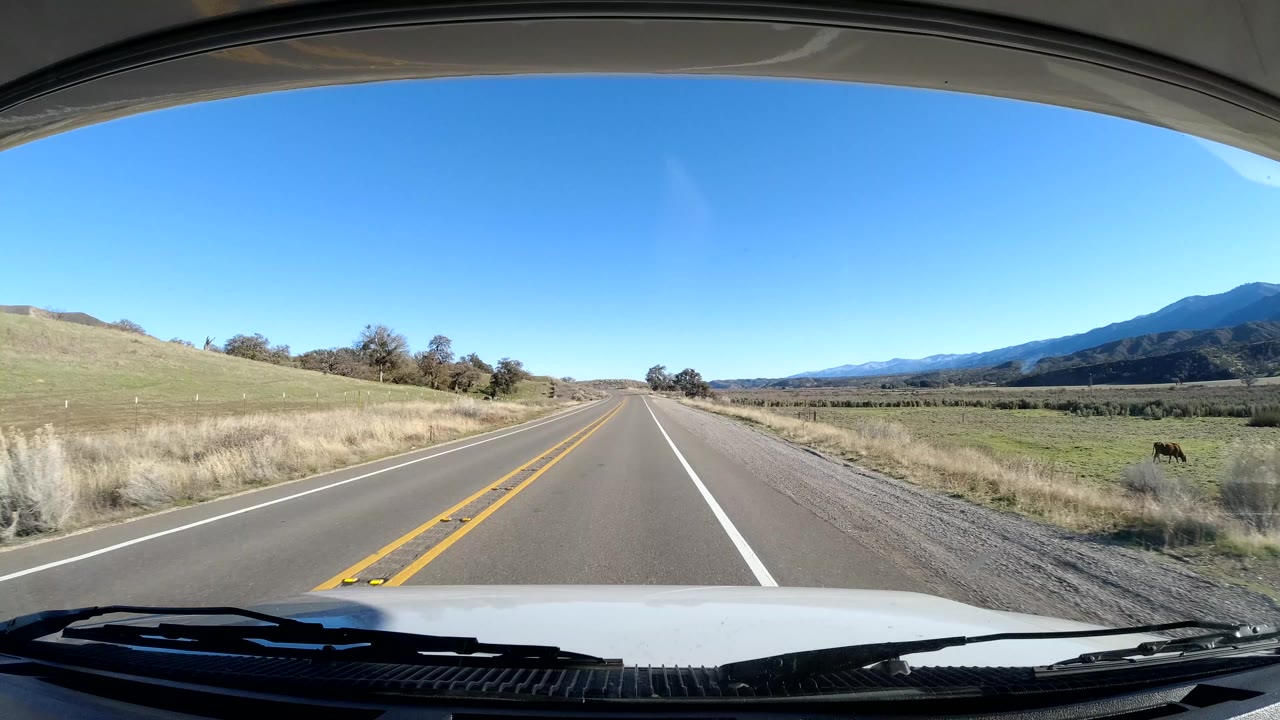 Central Coast California driving view at the start of the clip, showing a two-lane road, yellow center lines, grassy shoulders, distant mountains, vehicle hood, and no visible people.