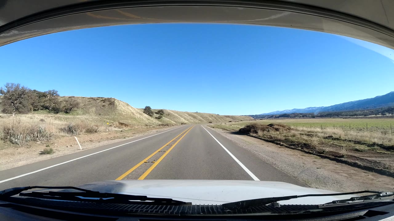 Central Coast California driving frame with a dirt hillside close to the right shoulder, dry grass, road markings, distant fields, mountains, windshield edge, and no visible people.