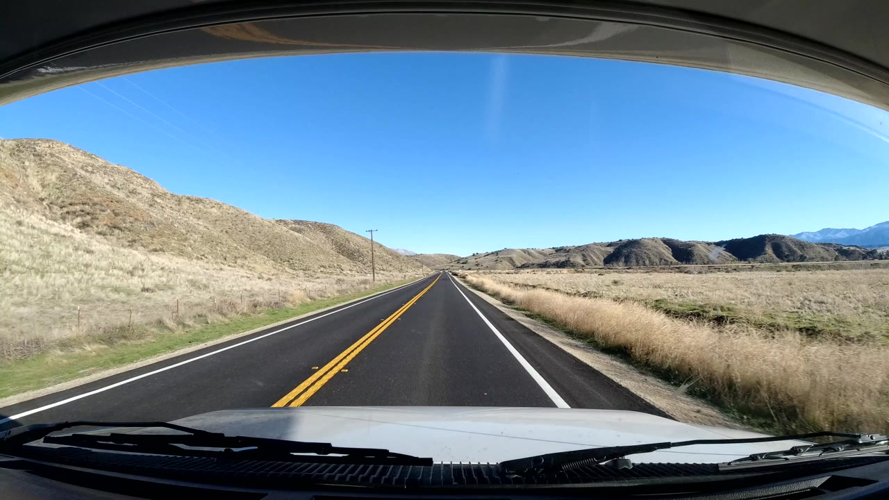 Forward road view in Coast California with a two-lane asphalt road, dry hills on both sides, blue sky, the vehicle hood at the bottom, and no visible people.
