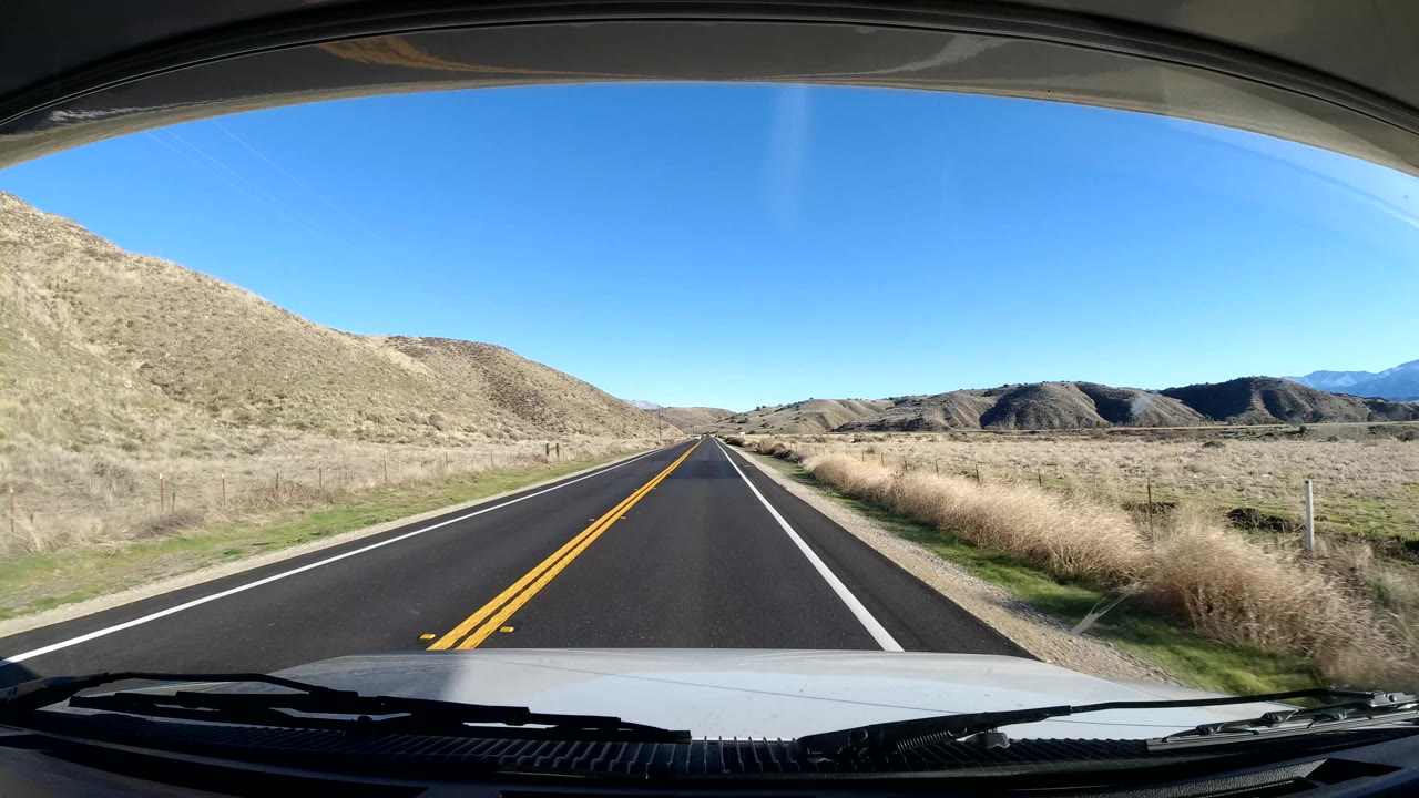 Straight rural road with dry grass and low hills ahead.