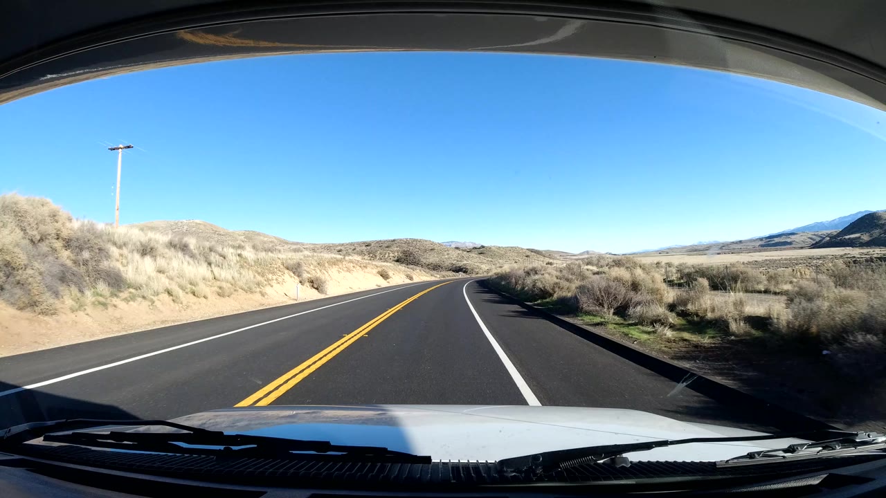 Forward vehicle view in Coast California as the road curves right past dry brush, sandy shoulder, rolling hills, a utility pole, and no visible people.