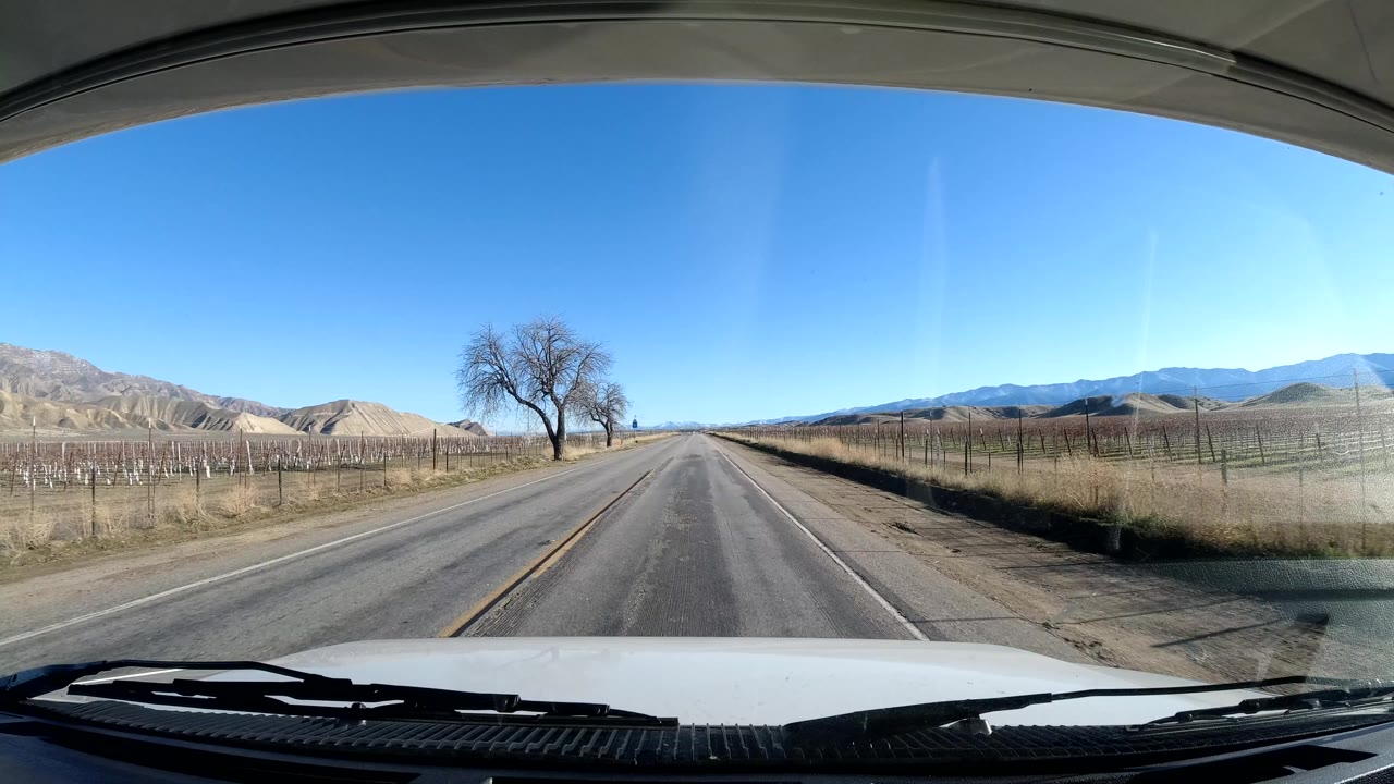 Opening frame of a Coast California rural road seen from inside a vehicle, with the hood and wipers at the bottom, fenced farmland on both sides, bare trees, distant hills, blue sky, and no visible people.