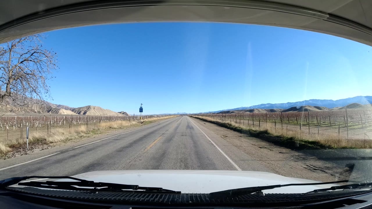 A Coast California roadway continues straight ahead from the windshield view, with a small roadside sign, wire fencing, dry agricultural fields, pale hills, distant mountains, and no visible people.