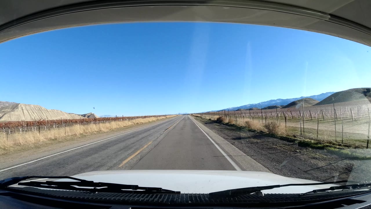 The vehicle view shows an empty Coast California two-lane road bordered by fenced farmland and dry shoulders, with layered hills in the background, a clear blue sky, and no visible people.