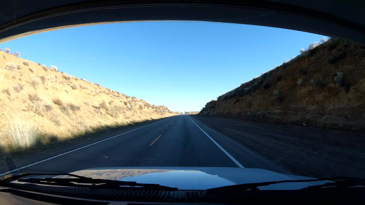 Dashboard-mounted view of a Central Coast California road entering a cut between dry hills, with the vehicle hood at the bottom and no visible people.