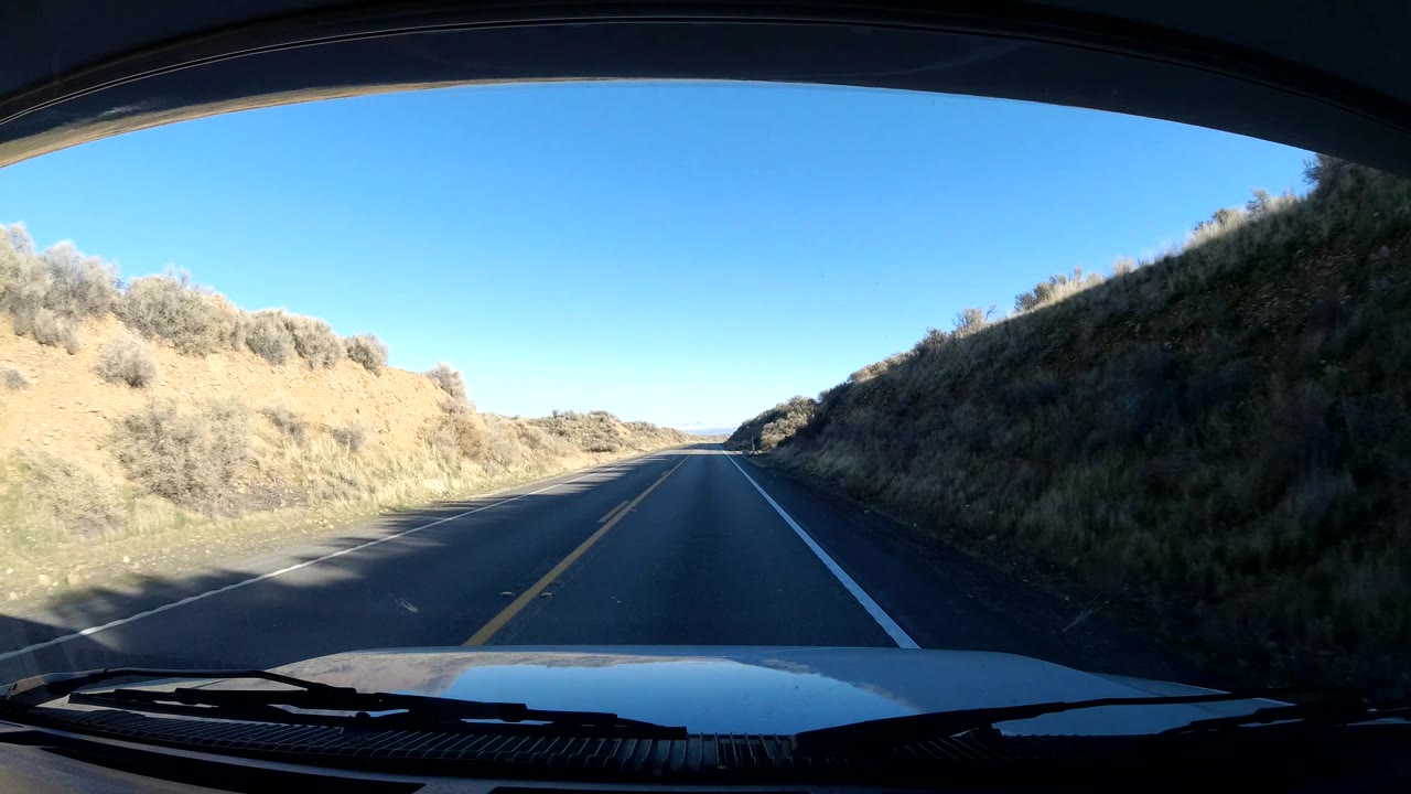 Dash-level Central Coast California road view with a shadowed embankment on the right, sunlit dry brush on the left, and no visible people.