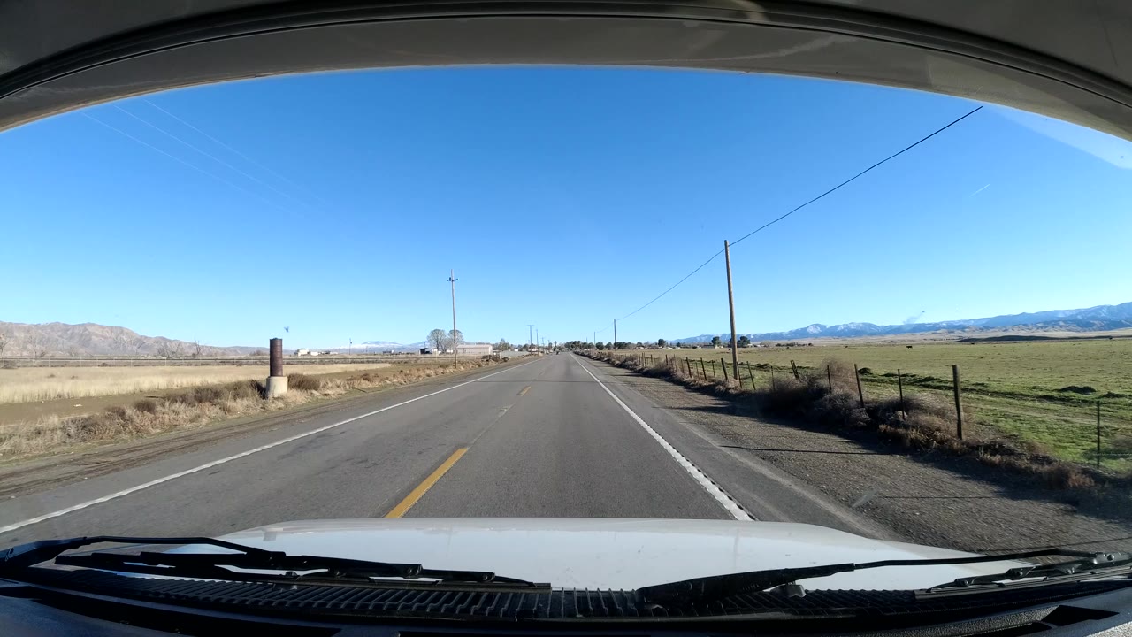 Dashboard view in Central Coast California passing open fields with fence posts, utility lines, and a low mountain range under clear sky, with no visible people.