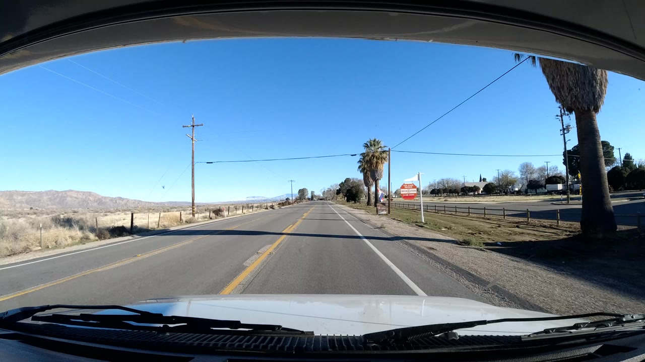 Dashboard view in Central Coast California approaching a roadside area with palm trees, utility poles, fencing, and a red sign on the right, with no visible people.