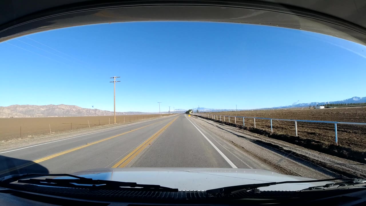 Straight rural road beside tilled fields with mountains in the distance.