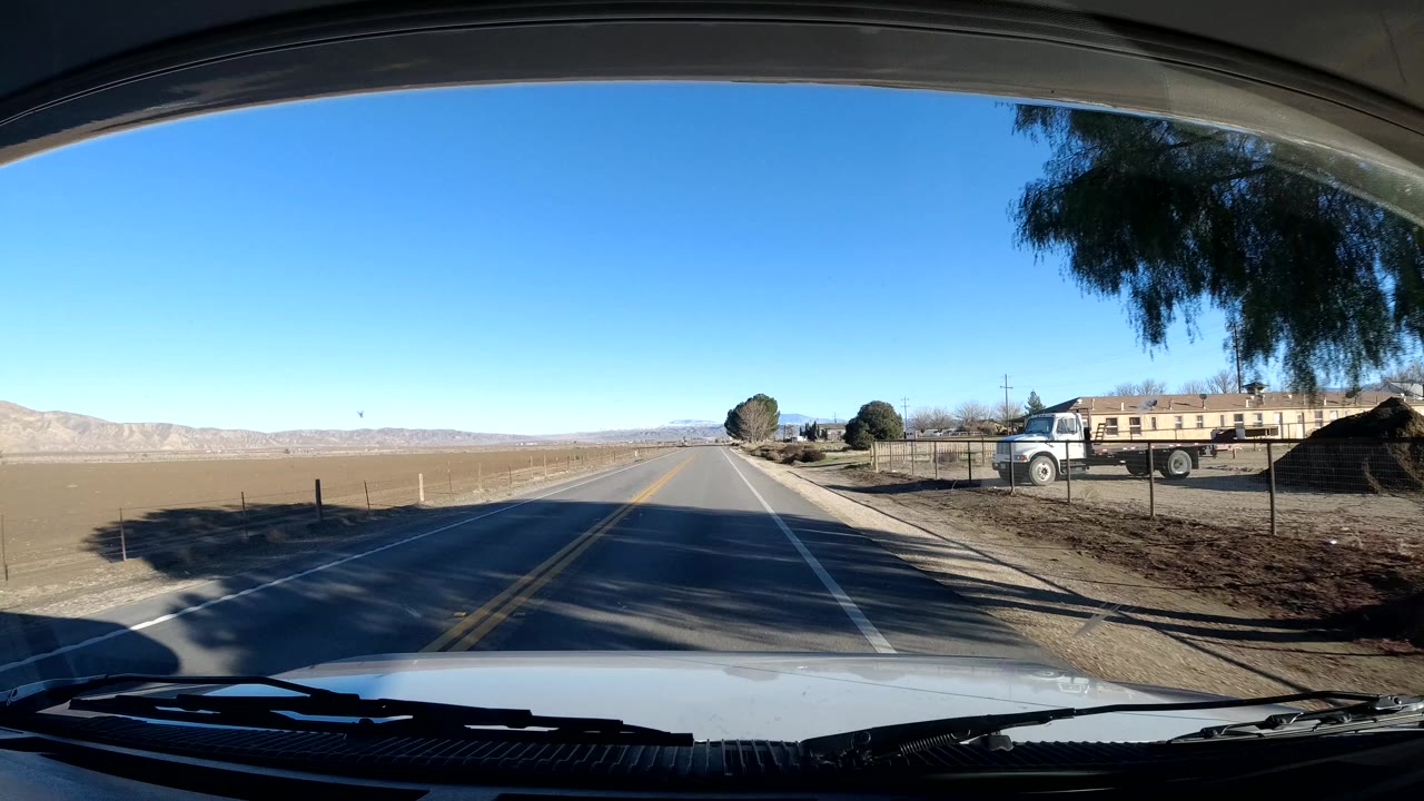 Windshield view from a vehicle on a Coast California road, with hood and wipers in the foreground, dry fields, a roadside building and truck, distant mountains, blue sky, and no visible people.
