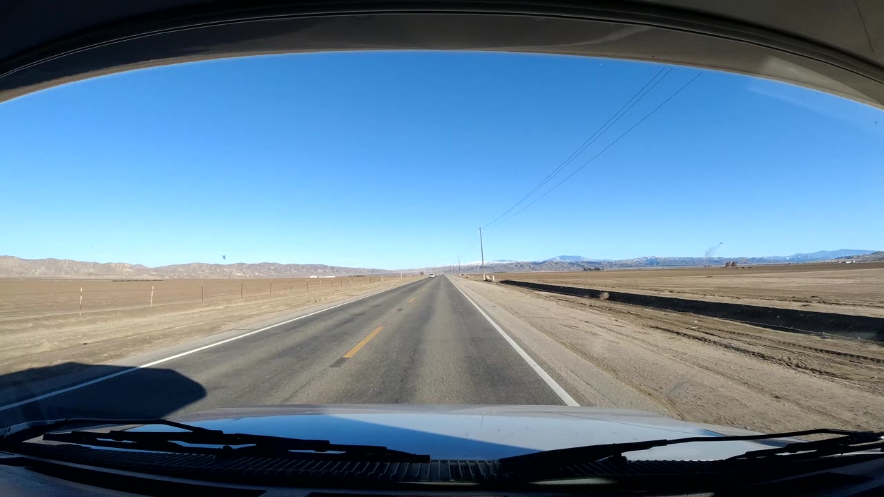 Straight rural highway with open fields and low mountains under blue sky.