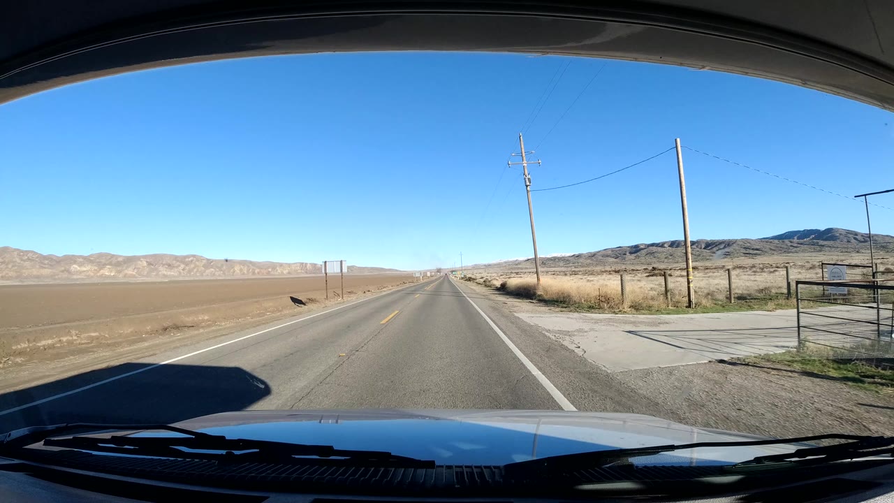 Vehicle windshield view on a rural Coast California road, with the hood and wipers in the foreground, utility poles beside dry fields, low hills ahead, and no visible people.
