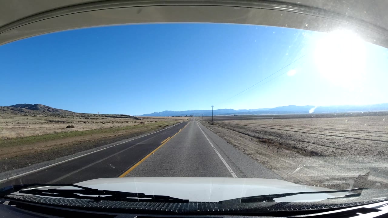 Coast California road view from inside a vehicle, with bright sun glare on the upper right, open fields on both sides, distant mountains, and no visible people.