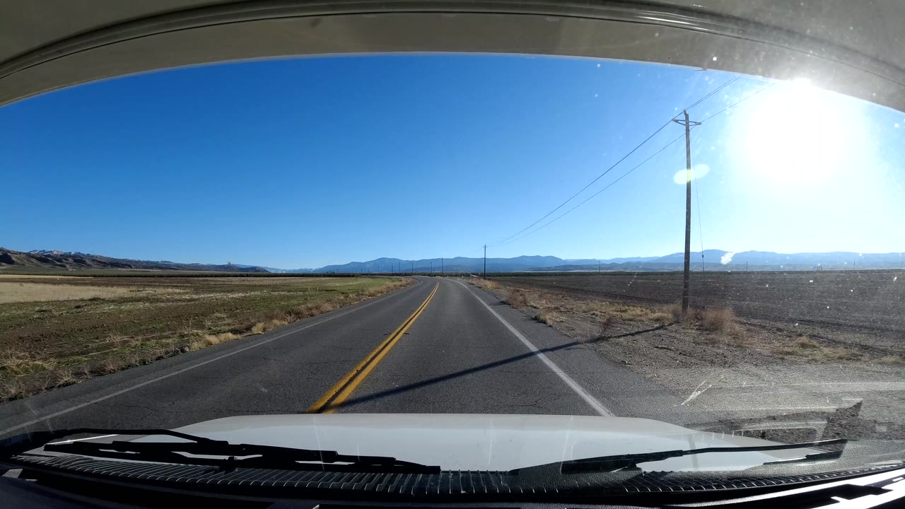 Curving rural road with a roadside pole, fields, and mountains in bright daylight.