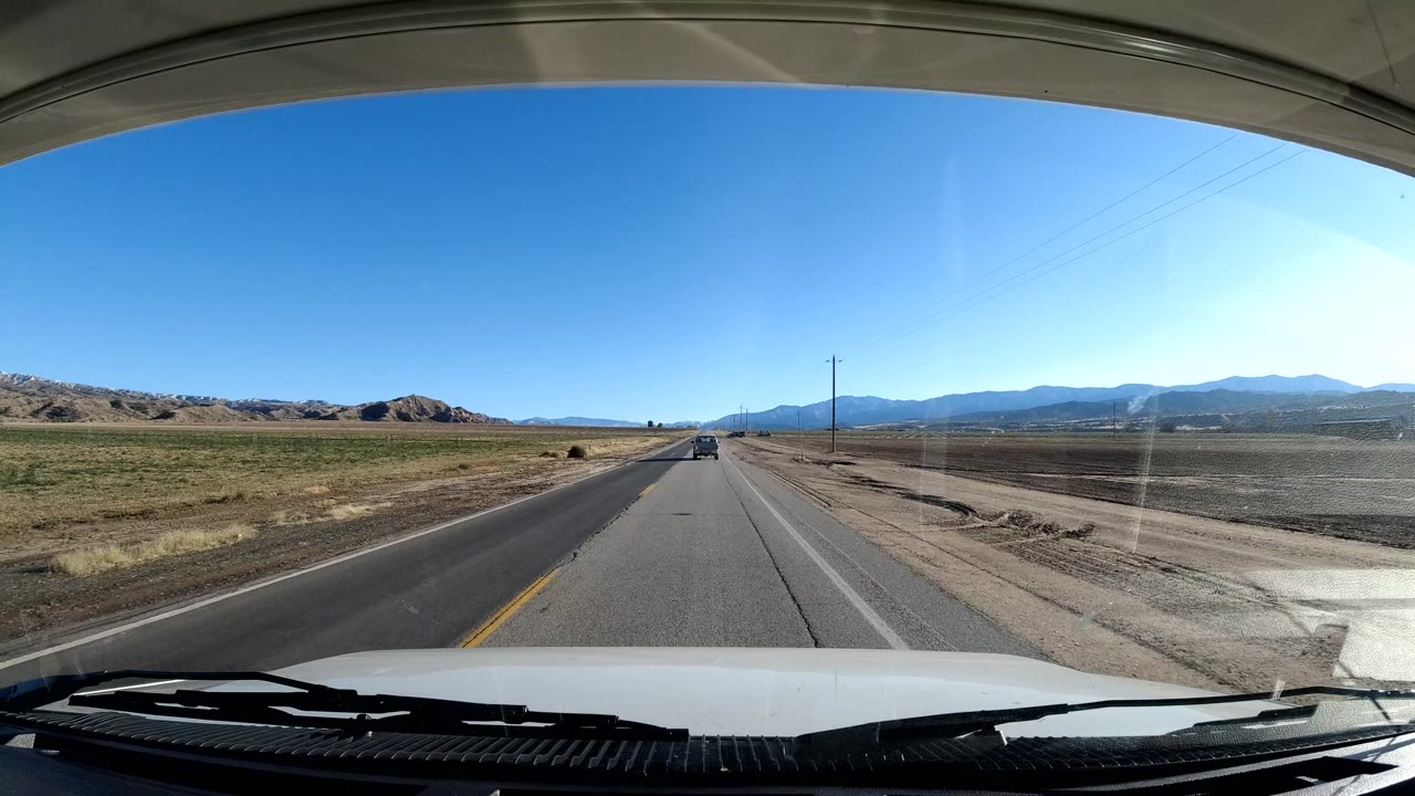 A Coast California road view through a vehicle windshield shows a straight two-lane road, open fields, distant mountains, blue sky, and no visible people.