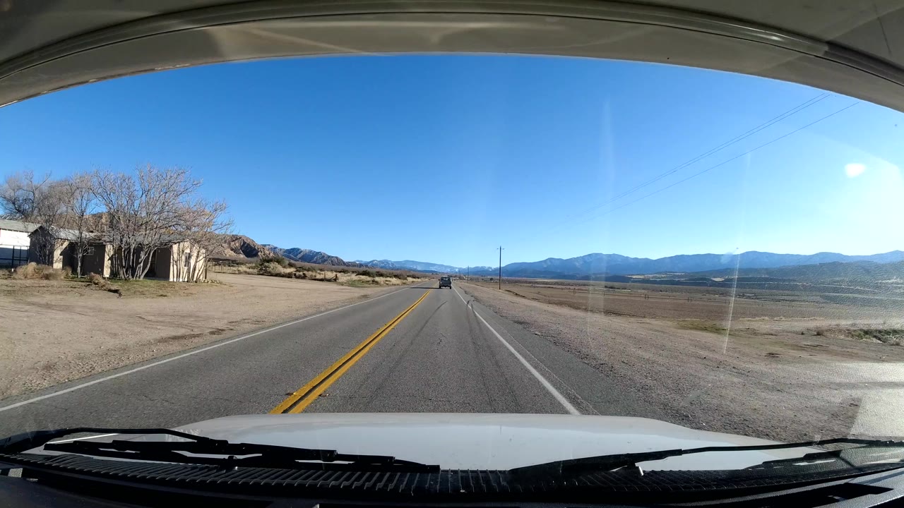 The Coast California vehicle POV passes a small roadside structure and bare trees on the left, with a two-lane road, fields, mountains, blue sky, and no visible people.