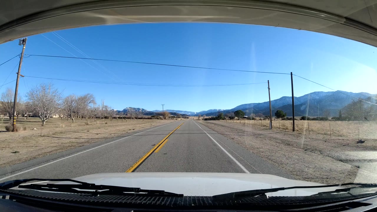 Dash-level Coast California road view with bare trees to the left, roadside poles, dry open land, mountains on the horizon, and no visible people.