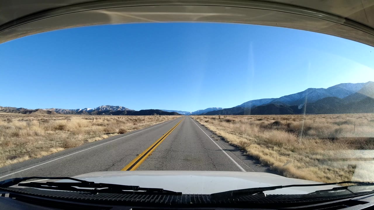 A forward vehicle view in Coast California shows an empty road through dry grassland, mountains along the right side, clear blue sky, and no visible people.