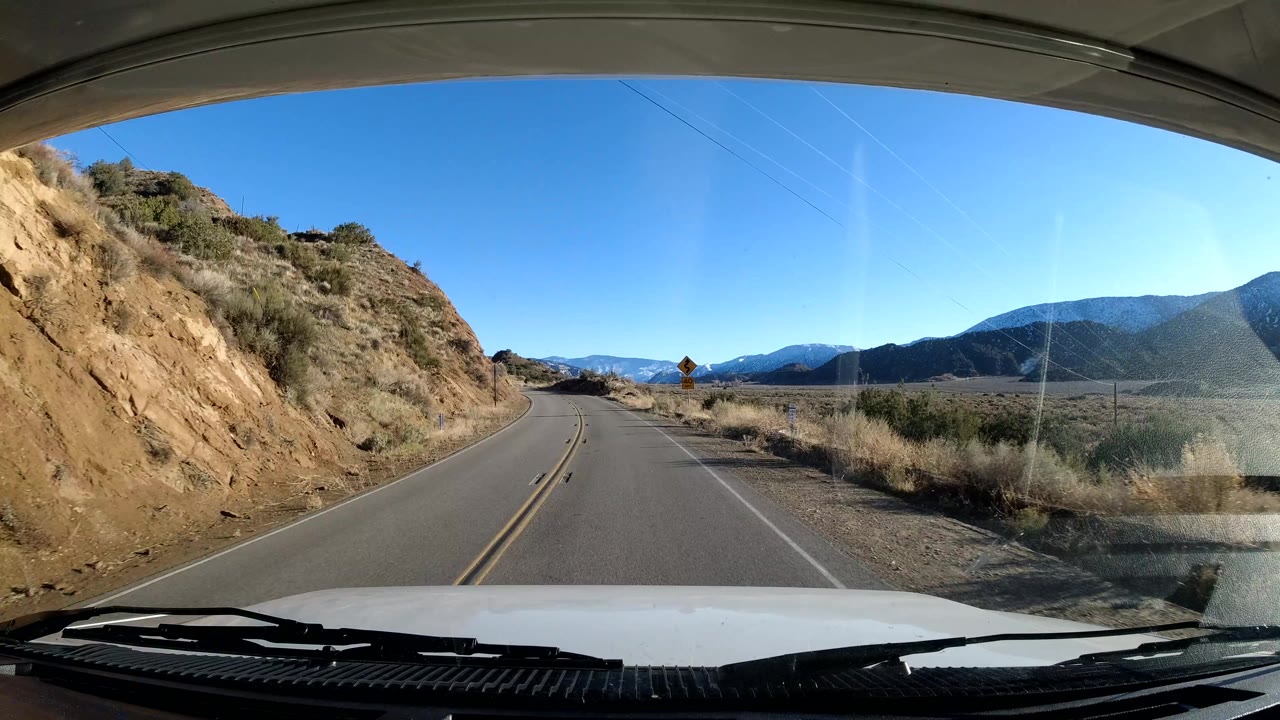 The Coast California drive reaches a curving road beside a cut hillside, with dry brush, mountains beyond the lane, windshield reflections, and no visible people.