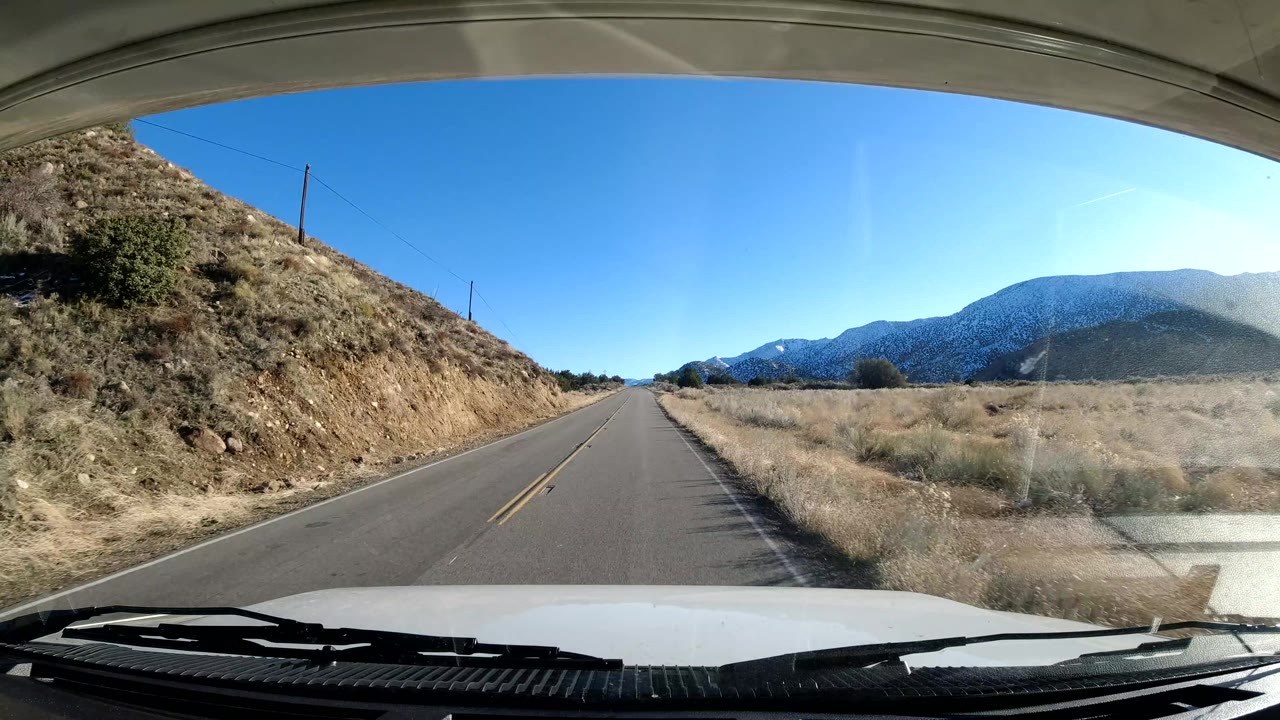 Vehicle-front view of a two-lane road in Coast California, with a dry hillside on the left, open field on the right, distant mountains, blue sky, and no visible people.